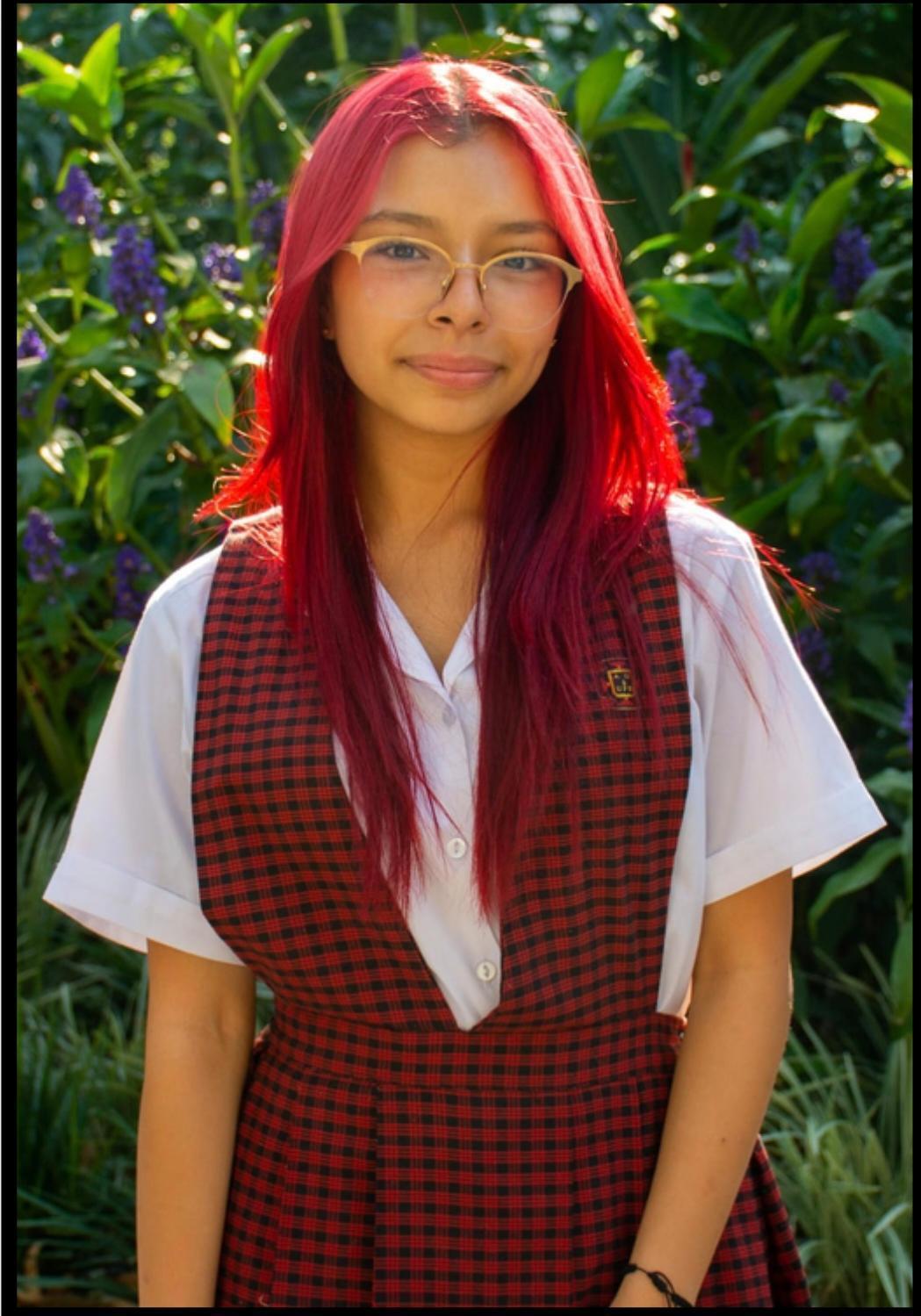 Estudiante con uniforme escolar rojo y blanco posando al aire libre frente a un jardín con flores y vegetación.