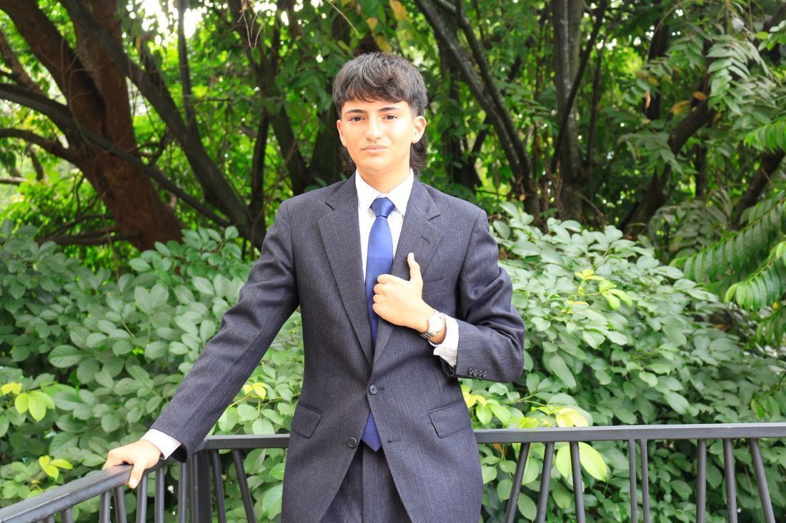 Retrato de Camilo Puerta vestido con traje formal oscuro y corbata azul. Se encuentra apoyado sobre una baranda en un espacio al aire libre rodeado de abundante vegetación. La fotografía transmite una imagen profesional 