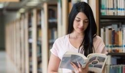 mujer con la cabeza baja leyendo un libro. De fondo se ven las estantería de una biblioteca